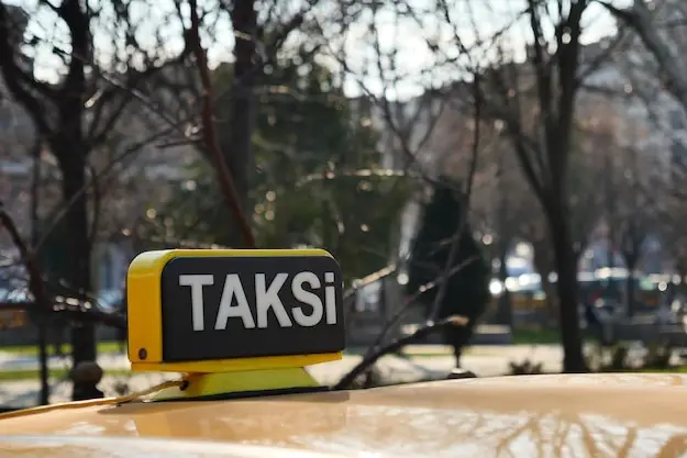 Ağaçlık bir yolda park hâlindeki sarı ticari taksinin çatısındaki TAKSİ tabelası